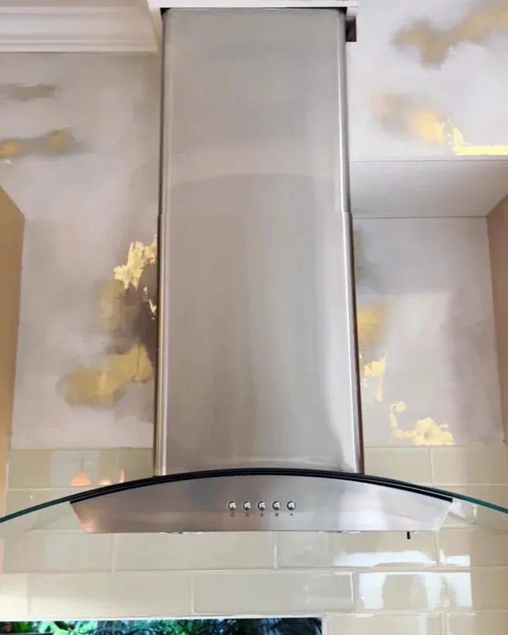 Stainless kitchen range hood against a wall of soft gray and gold leaf